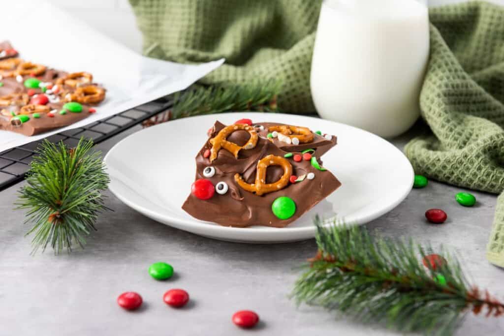 Piece of milk chocolate reindeer bark Christmas treats on a plate surrounded by fir branches and a glass bottle of milk. 