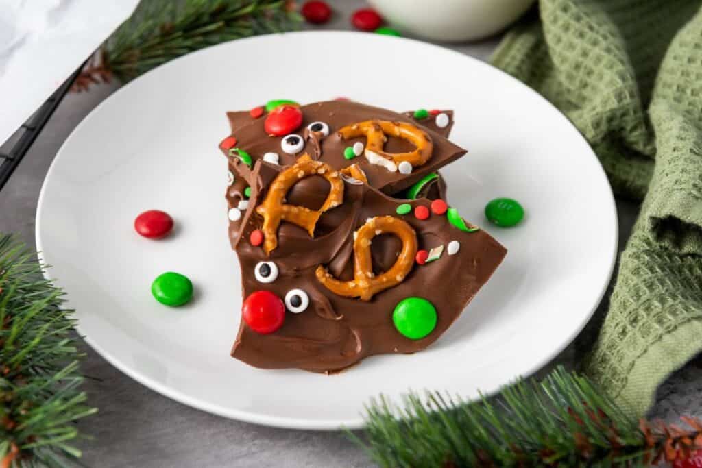 Pieces of chocolate bark candy treat with little reindeer faces pressed in, made out of candy eyes, pretzel antlers and M&M noses.