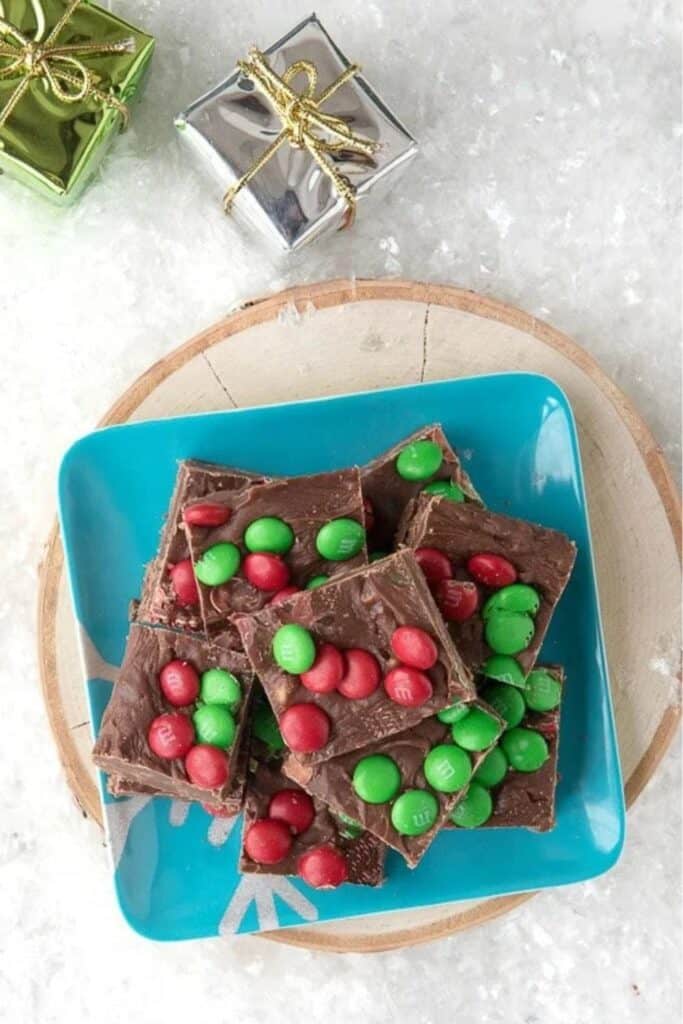 A festive blue plate stacked with squares of chocolate toffee holiday fudge, topped with red and green M&M candies. The fudge sits on a wooden tray surrounded by faux snow and small wrapped presents.