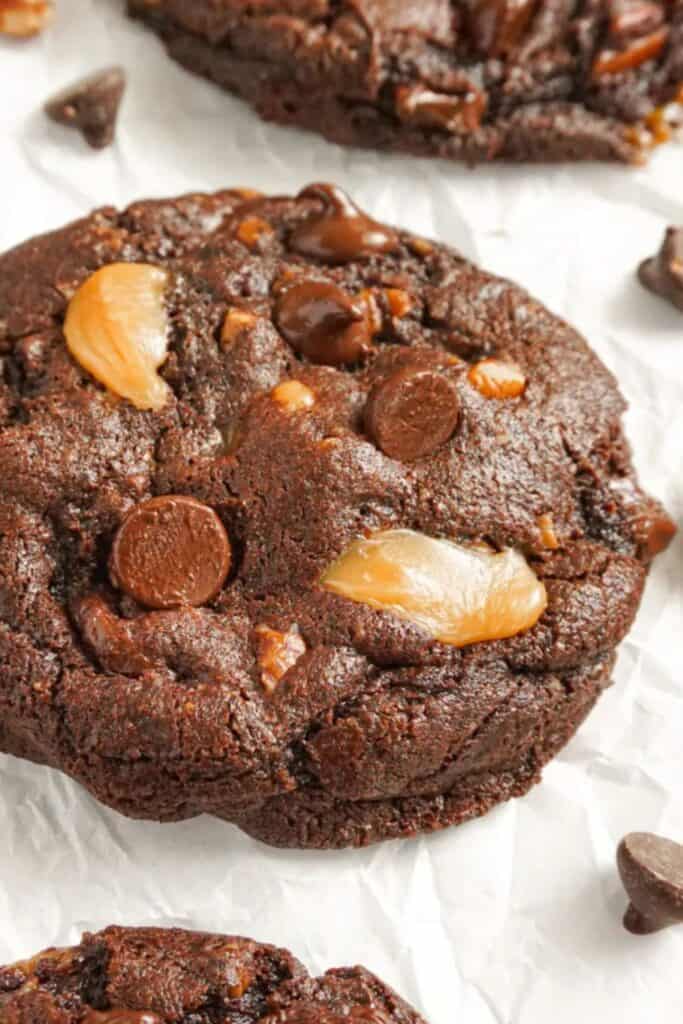 Close-up of a rich, fudgy chocolate turtle cookie loaded with melted caramel chunks and chocolate chips, resting on crinkled parchment paper. The gooey textures and deep cocoa color make it a decadent and easy Christmas cookie