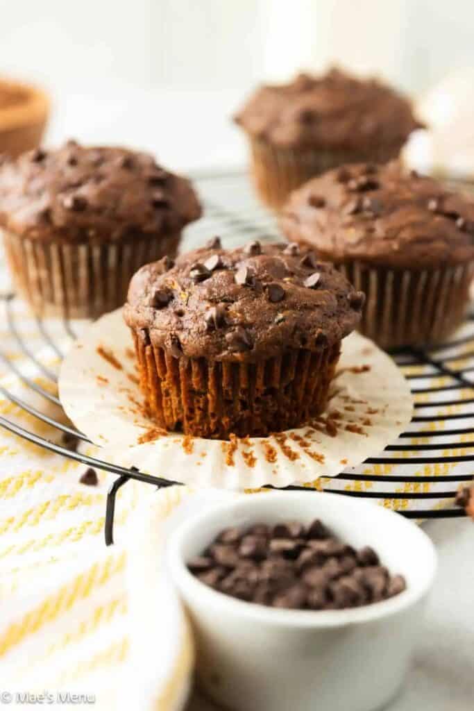 Moist chocolate zucchini muffin with a cracked top and mini chocolate chips, sitting on an unwrapped liner atop a cooling rack. Surrounded by more muffins and a small bowl of chocolate chips.