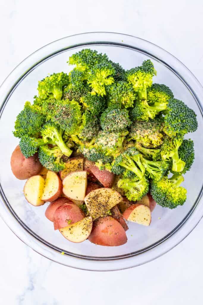 Overhead view of a glass bowl filled with raw broccoli florets and halved baby red potatoes, seasoned with black pepper and spices. The vegetables are prepped and ready to be roasted for an easy sheet pan dinner.