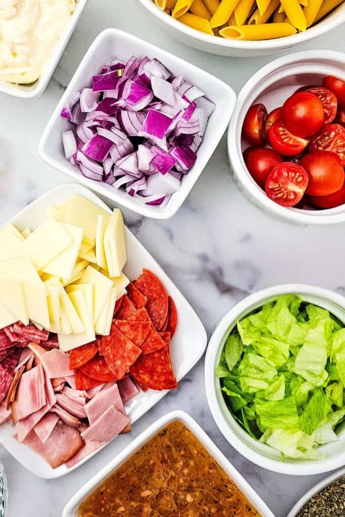  Overhead shot of prepped ingredients for a cold sub pasta salad, including diced red onion, halved cherry tomatoes, shredded lettuce, sliced provolone cheese, ham, pepperoni, salami.