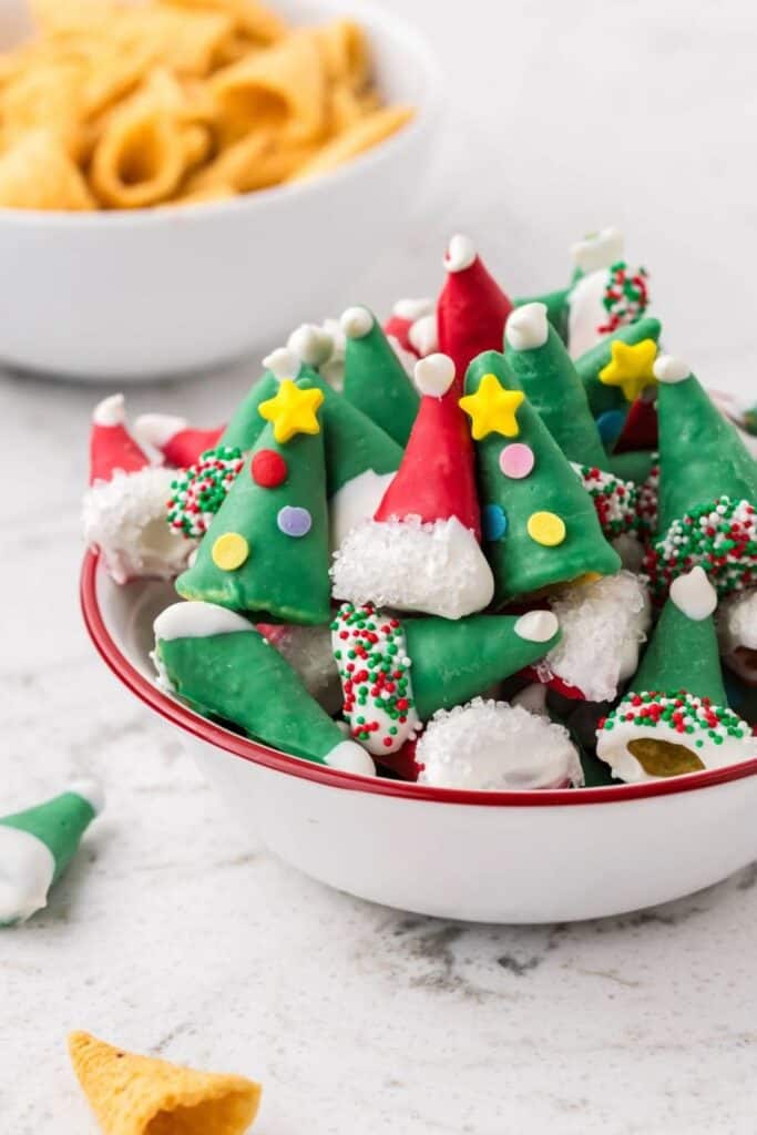 A bowl of decorated Christmas Bugles made to look like mini Christmas trees and Santa hats, dipped in red and green candy coating with festive sprinkles, white chocolate trim, and candy embellishments, served as a whimsical treat for holiday snacking.