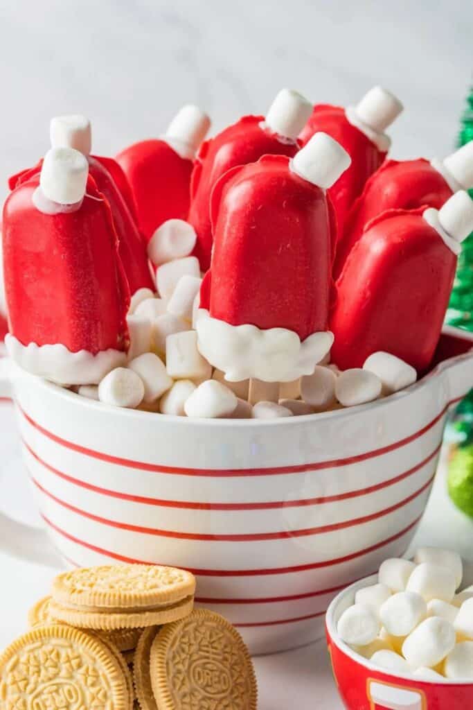 Red and white Christmas Cakesicles shaped like Santa hats, dipped in red candy coating with a white chocolate trim and mini marshmallow toppers. Displayed in a striped bowl filled with marshmallows, with golden Oreos and holiday decorations nearby.