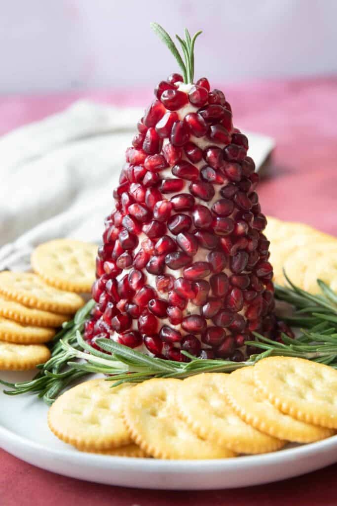 Christmas cheese ball shaped like a pine cone and covered with bright red pomegranate seeds. Fresh rosemary sprigs decorate the top and base with round golden crackers arranged around the plate for serving.