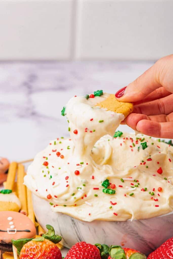 A hand dips a round cookie into a bowl of creamy white Christmas cookie dip topped with red, green, and white holiday sprinkles, with strawberries, cookies, and pretzel sticks arranged around the bowl.