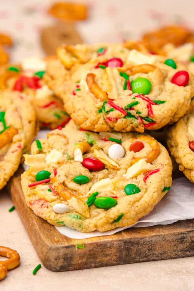 Close up of colorful, easy Christmas cookies on a wooden board, featuring red and green M&Ms, white chocolate chunks, pretzel pieces, potato chips, and red and green sprinkles baked into golden, chewy cookies.