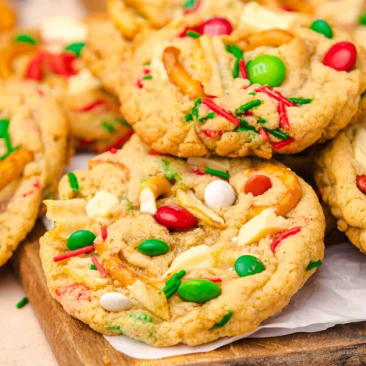 Close up of colorful, easy Christmas cookies on a wooden board, featuring red and green M&Ms, white chocolate chunks, pretzel pieces, potato chips, and red and green sprinkles baked into golden, chewy cookies.