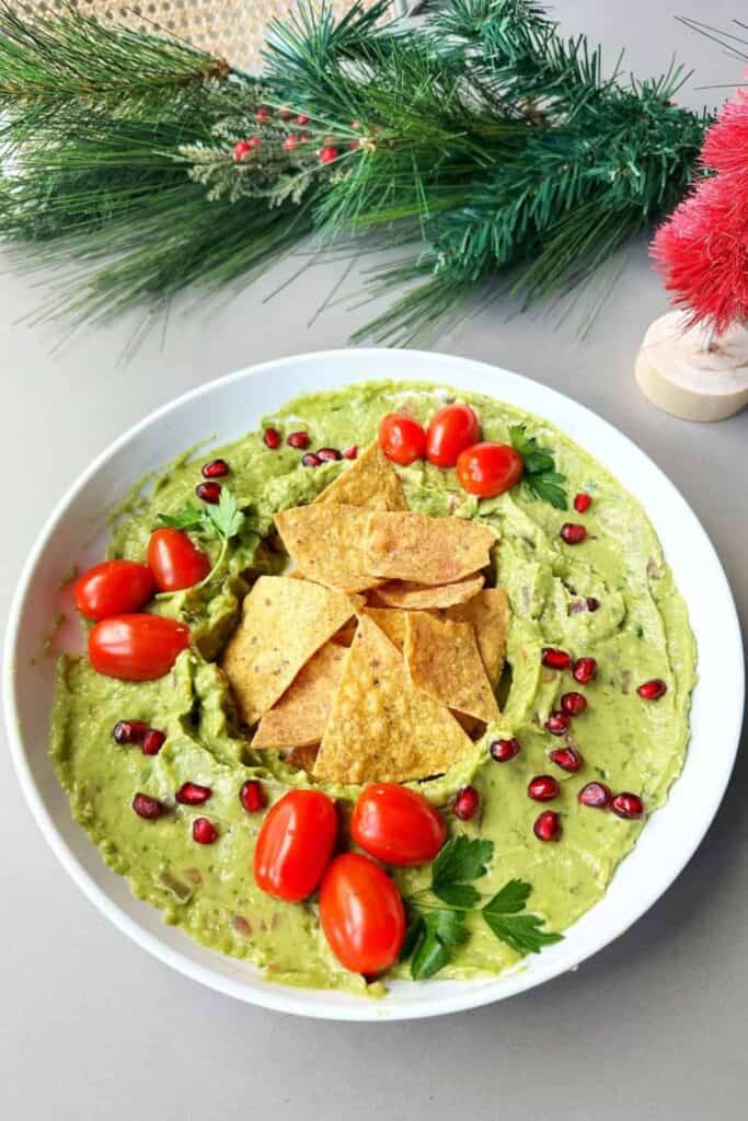 A festive bowl of guacamole garnished with grape tomatoes, pomegranate seeds, and parsley, arranged in a holiday wreath shape with tortilla chips in the center, making it a colorful and savory movie night snack.