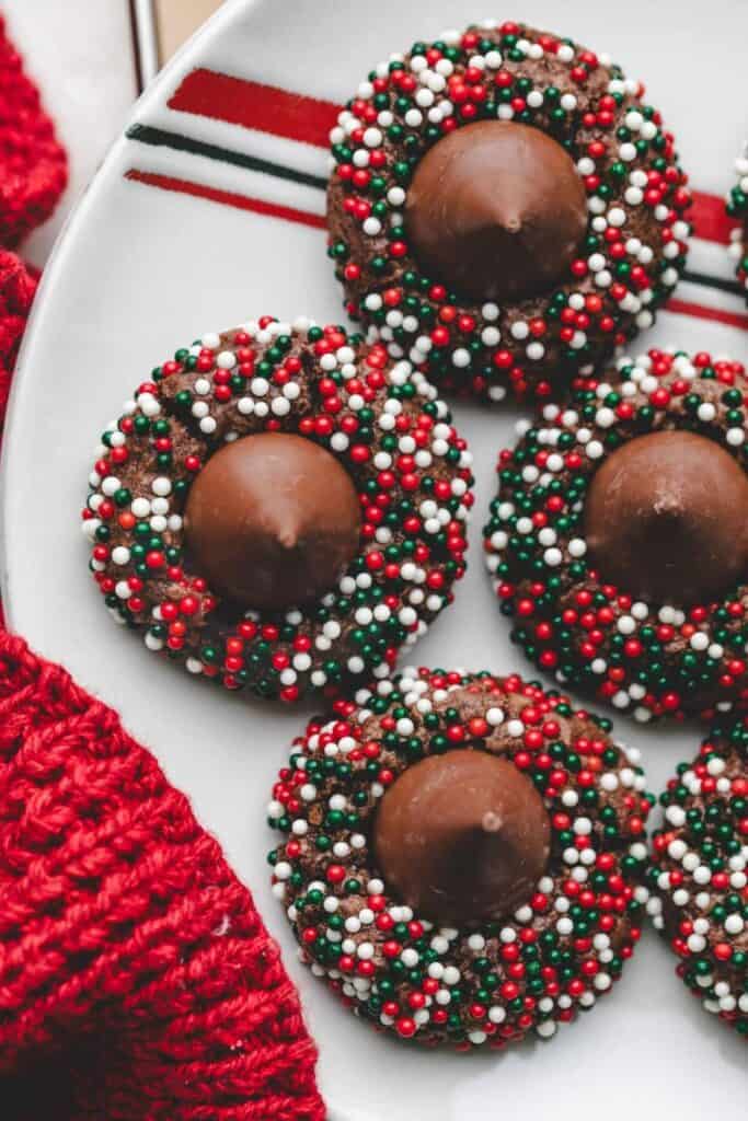 Plate of chocolate kiss cookies decorated with festive red, white, and green sprinkles, each topped with a milk chocolate Hershey’s Kiss. The cookies sit beside a red knit cloth.