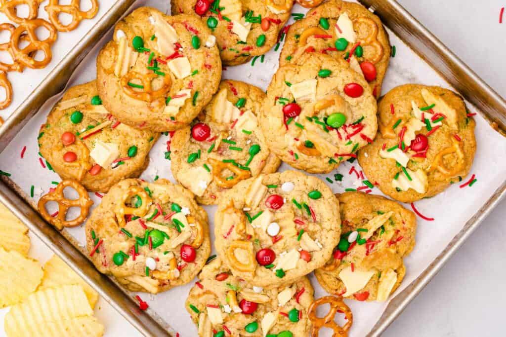 Overhead view of a baking sheet filled with Christmas Kitchen Sink cookies topped with red and green M&Ms, pretzels, sprinkles, white chocolate chunks, and crushed potato chips, surrounded by extra pretzels and chips on the side.