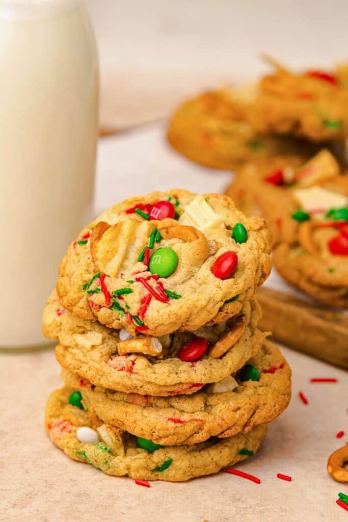 Stack of Christmas M&M cookies filled with red and green M&Ms, white chocolate chunks, pretzels, potato chips, and festive sprinkles, with a glass of milk in the background and more cookies slightly blurred behind.