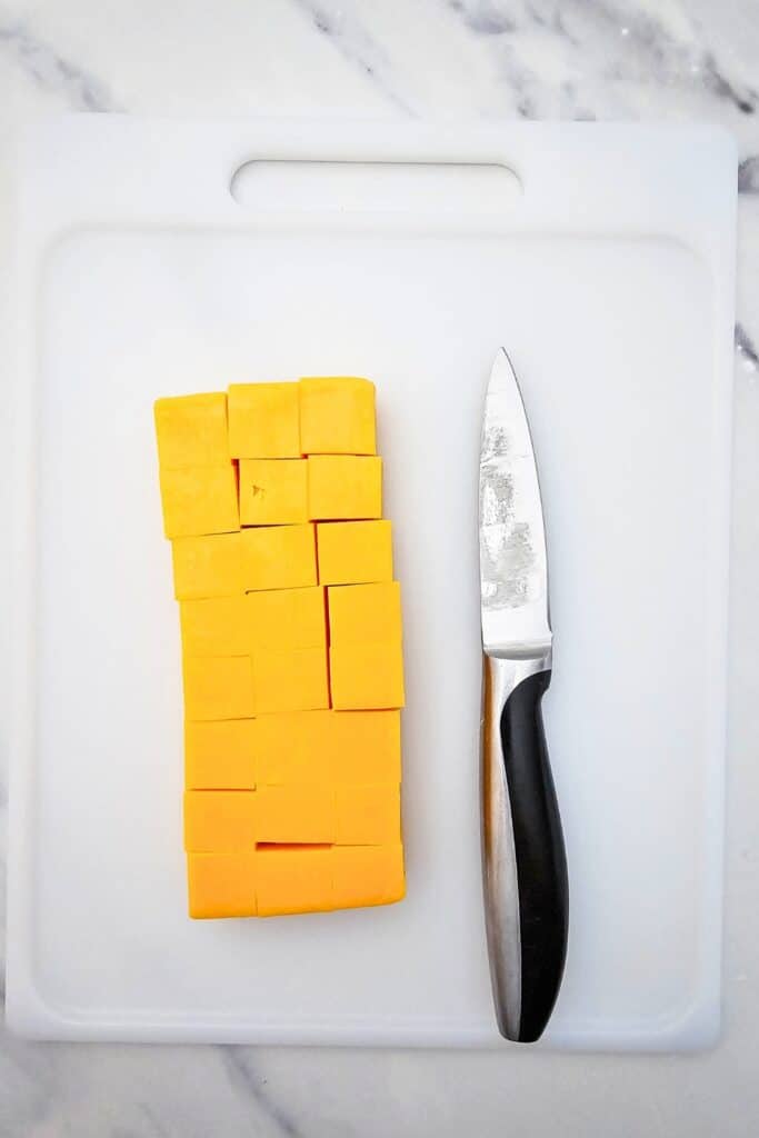 Cubed cheddar cheese arranged beside a kitchen knife on a white cutting board, ready to add to a Christmas party fruit tray with fresh grapes, raspberries, and rosemary.