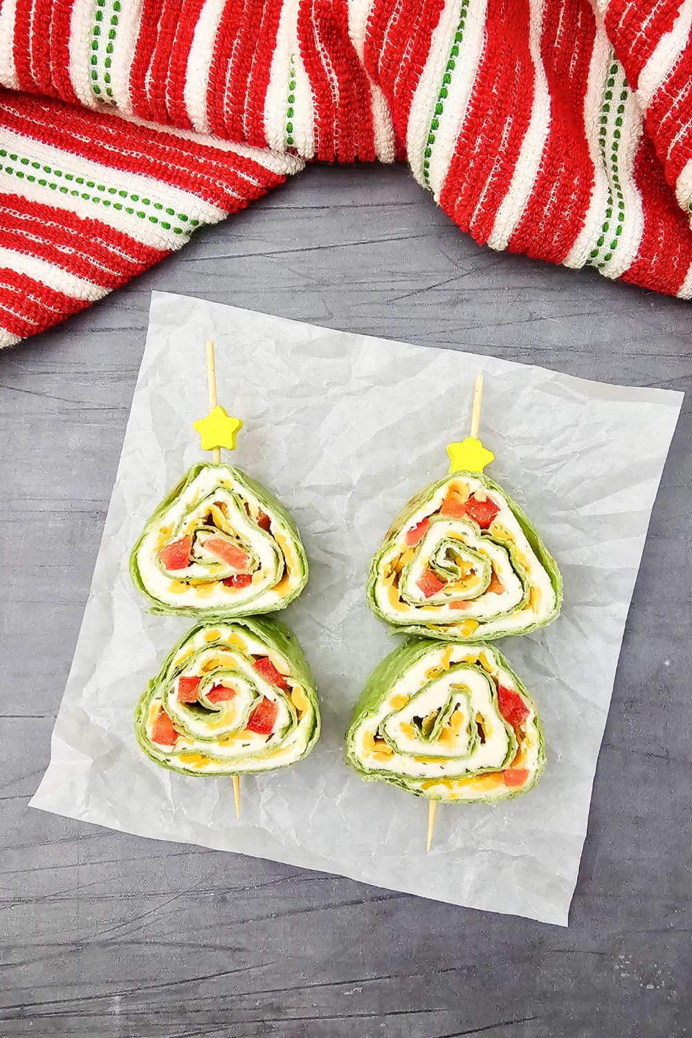 Two Christmas tortilla pinwheels on skewers, shaped like festive trees with layers of spinach wraps, cream cheese, cheddar, and red peppers, displayed on parchment paper with a red and white holiday towel in the background. A fun and colorful holiday party finger food idea.