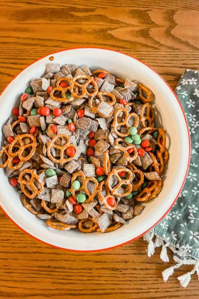 A large bowl of Christmas puppy chow made with chocolate-covered Chex cereal coated in powdered sugar, mixed with mini pretzels and red and green M&Ms for a festive, sweet-and-salty holiday snack.