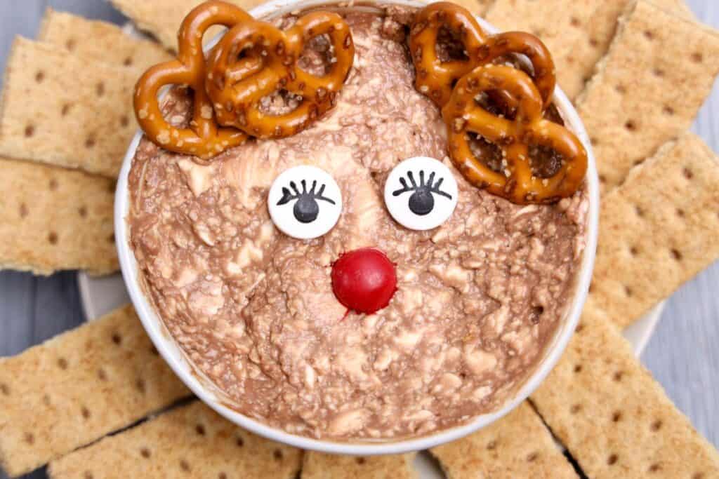 Rudolph-inspired Christmas dip in a bowl, styled to look like a reindeer with pretzel antlers, candy eyes with drawn-on lashes, and a red candy nose. The creamy, chocolatey dip is surrounded by rectangular graham crackers arranged like a sunburst.