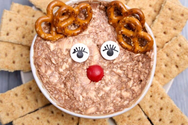 Rudolph-inspired Christmas dip in a bowl, styled to look like a reindeer with pretzel antlers, candy eyes with drawn-on lashes, and a red candy nose. The creamy, chocolatey dip is surrounded by rectangular graham crackers arranged like a sunburst.