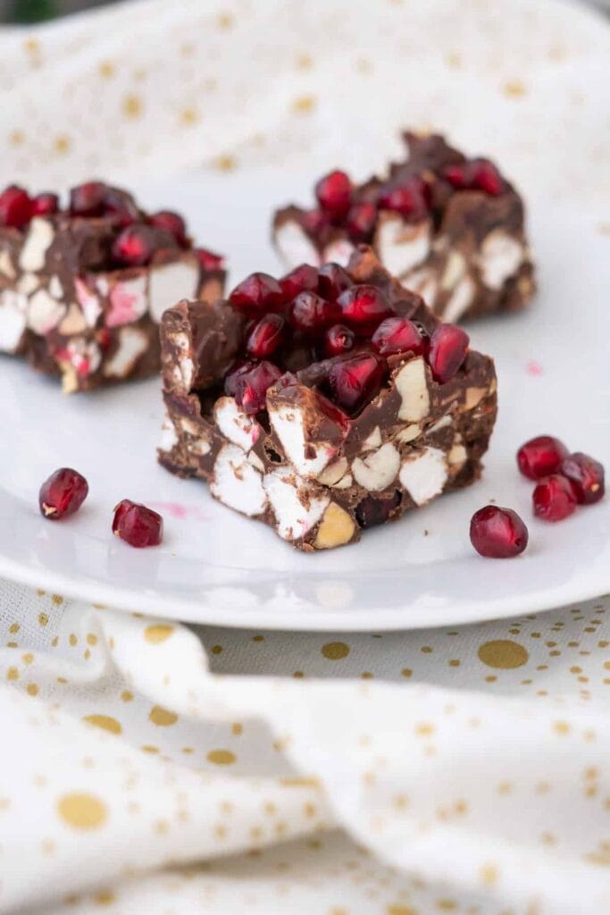 Christmas rocky road bars made with chocolate, mini marshmallows, nuts, and festive red pomegranate seeds on top. Served on a white plate with a gold polka dot napkin for a cheerful holiday dessert.
