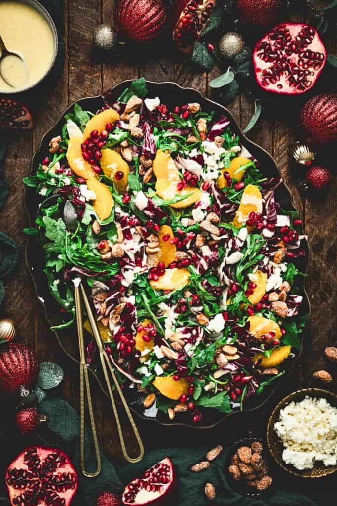 An overhead view of a festive Christmas salad potluck recipe with mixed greens, radicchio, orange slices, pomegranate seeds, crumbled cheese, and spiced nuts, surrounded by pomegranates and Christmas ornaments with a creamy dressing on the side.
