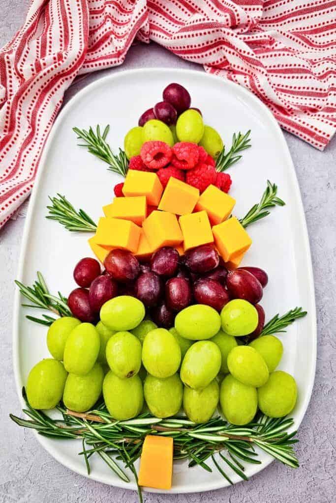 A Christmas themed charcuterie board arranged like a Christmas tree with layers of green and red grapes, fresh raspberries, cheddar cheese cubes, and rosemary branches on a white oval platter beside a red and white holiday towel.