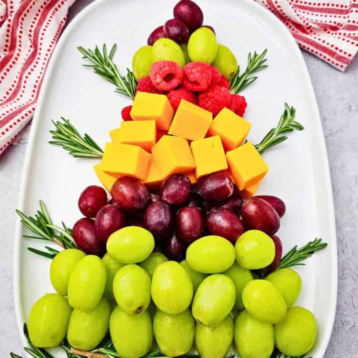 A Christmas themed charcuterie board arranged like a Christmas tree with layers of green and red grapes, fresh raspberries, cheddar cheese cubes, and rosemary branches on a white oval platter beside a red and white holiday towel.
