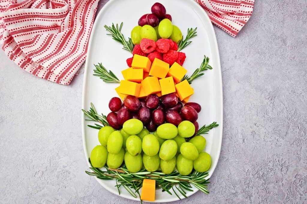 Christmas Tree Fruit Tray arranged on a white oval platter with layers of green and red grapes, fresh raspberries, cheddar cheese cubes, and rosemary sprigs, creating a colorful festive appetizer and snack for holiday gatherings.