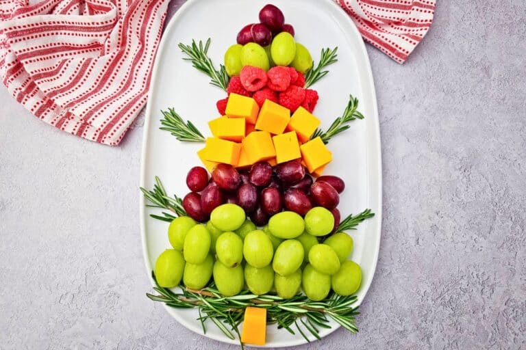Christmas Tree Fruit Tray arranged on a white oval platter with layers of green and red grapes, fresh raspberries, cheddar cheese cubes, and rosemary sprigs, creating a colorful festive appetizer and snack for holiday gatherings.