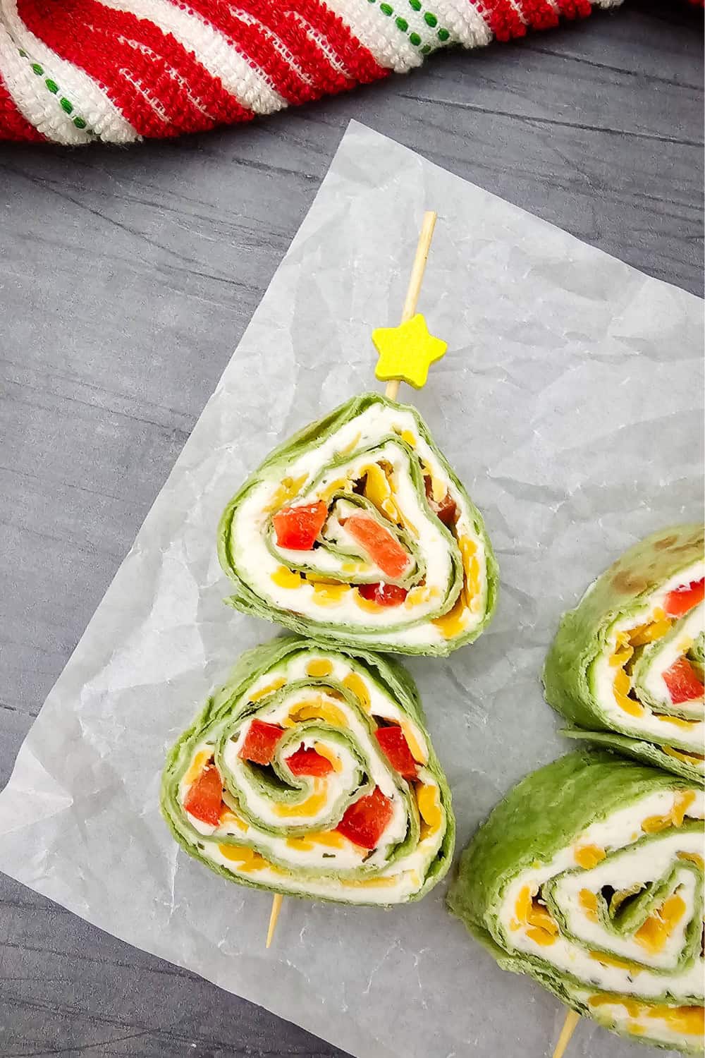Christmas tree shaped pinwheel appetizers made with green tortillas rolled with cream cheese and filled with red and yellow bell peppers. Two pinwheels are stacked on a wooden skewer to form a Christmas tree and topped with a yellow star.