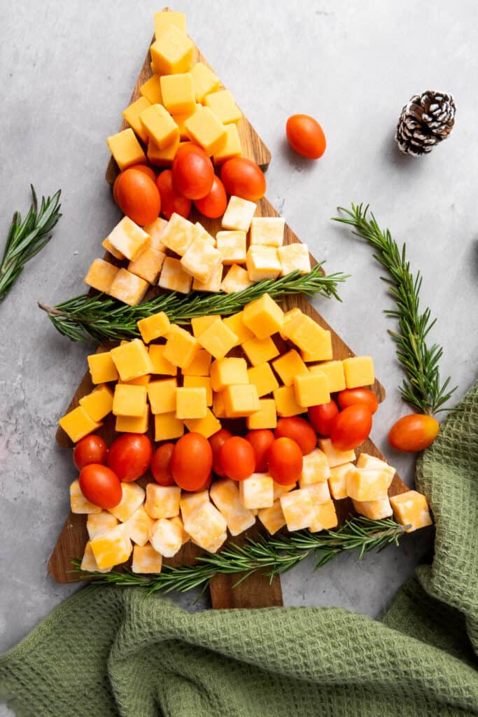 Christmas Tree shaped wooden cutting board with rows of alternating cheese cubes, cherry tomatoes and rosemary sprigs