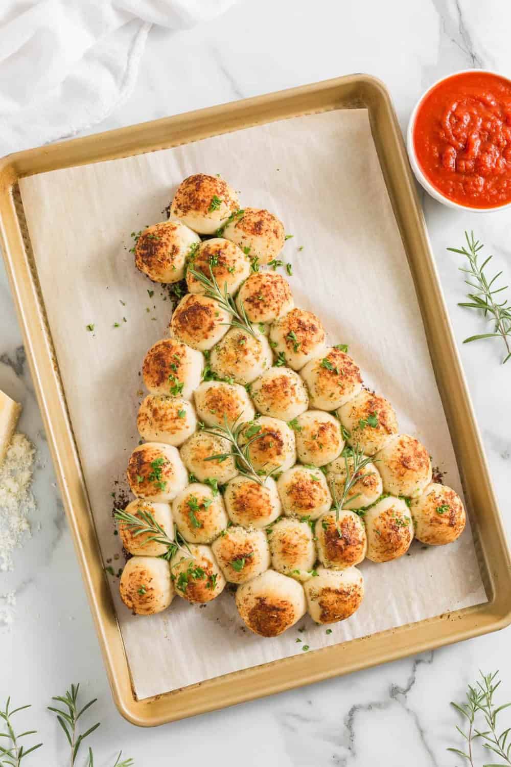 hristmas tree pull apart bread made with golden baked rolls arranged in a tree shape and garnished with fresh rosemary and parsley. A bowl of red marinara sauce sits alongside for dipping.
