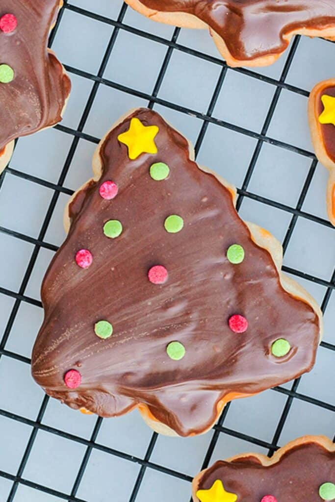 Christmas tree-shaped Twix cookie topped with smooth chocolate frosting, decorated with red and green candy dots and a yellow star sprinkle. The cookie sits on a black wire cooling rack.