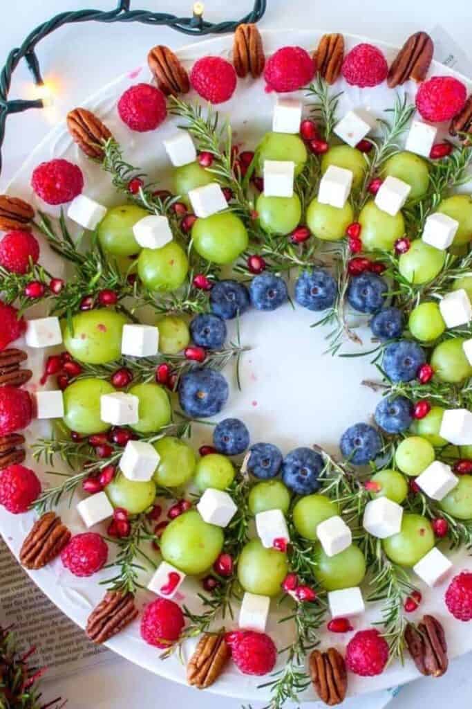 Christmas wreath fruit board arranged with green grapes, blueberries, raspberries, pomegranate seeds, pecans, cheese cubes, and fresh rosemary. The colorful ingredients form a festive edible wreath on a round white platter.