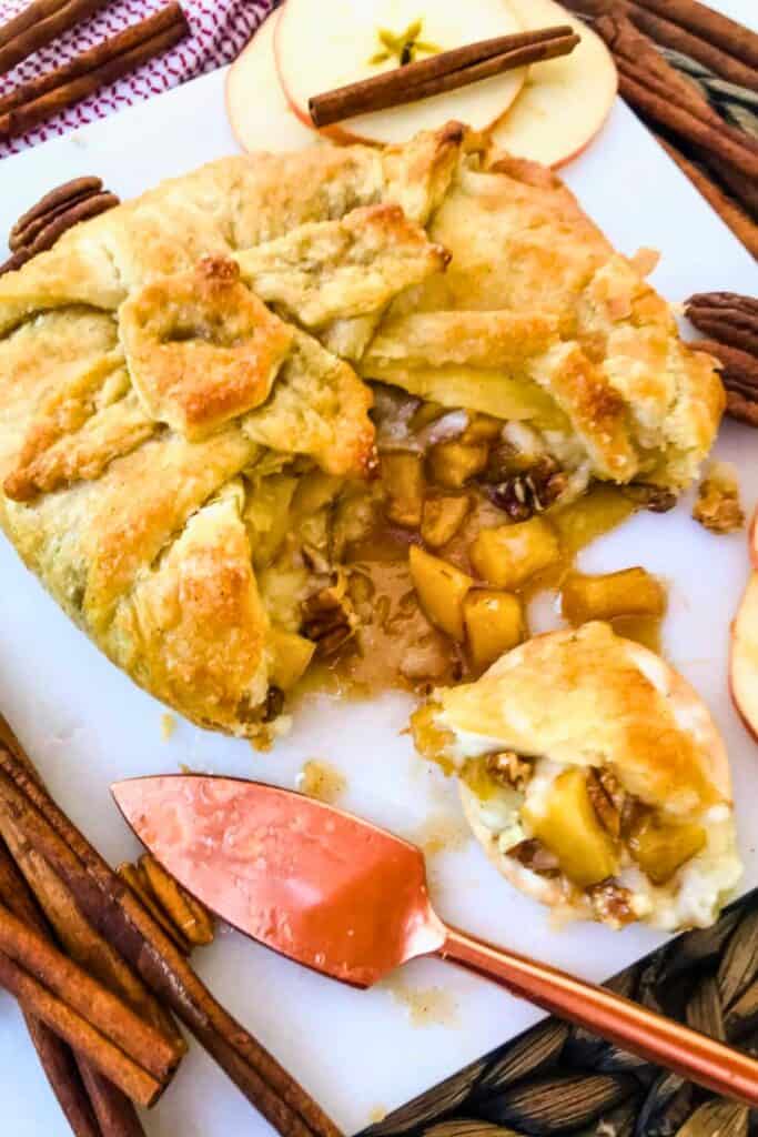 Cinnamon apple baked brie wrapped in golden puff pastry is cut open to reveal melted cheese, tender apple pieces, pecans, and a gooey cinnamon filling. A sliced portion sits beside the baked brie with fresh apple slices, cinnamon sticks, pecans, and a copper serving knife arranged around the platter.