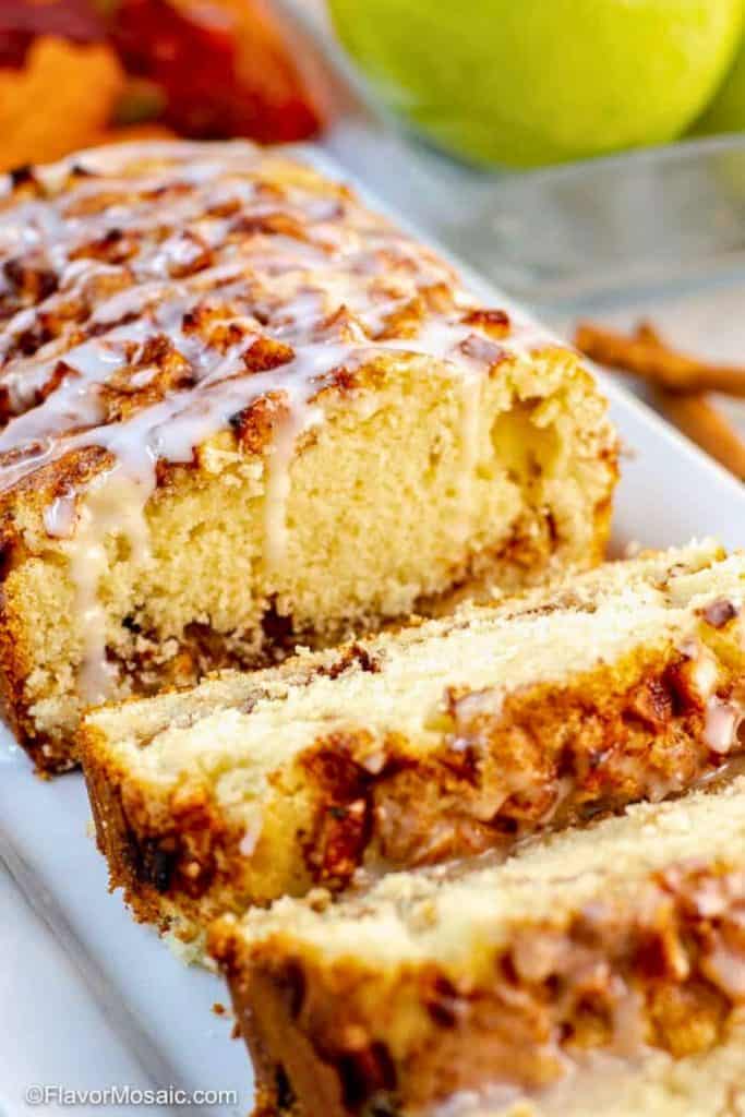 Close-up of a sliced apple cinnamon loaf cake topped with a light vanilla glaze, showing its moist, golden crumb and chunks of spiced apple and nuts throughout. Warm tones and whole apples in the background highlight its fall-inspired flavors.