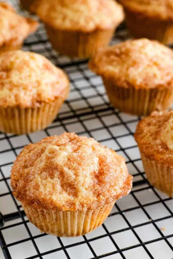 Freshly baked cinnamon muffins cooling on a wire rack with a golden brown crumb topping and sugar dusted surface. Multiple muffins are arranged in the background highlighting their soft texture and slightly domed tops.