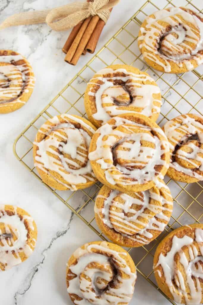 Overhead view of Cinnamon Roll Cookies with cinnamon sugar swirls and white icing drizzled over the tops, arranged on a gold cooling rack beside bundled cinnamon sticks.