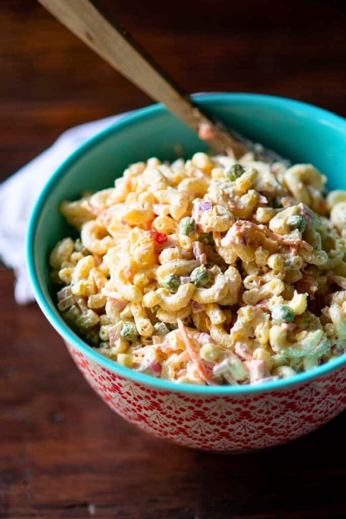 A vibrant turquoise and red patterned bowl filled with creamy macaroni salad, featuring elbow pasta, green peas, diced red onions, shredded carrots, and a rich mayonnaise-based dressing. A wooden spoon rests in the bowl, ready for serving.