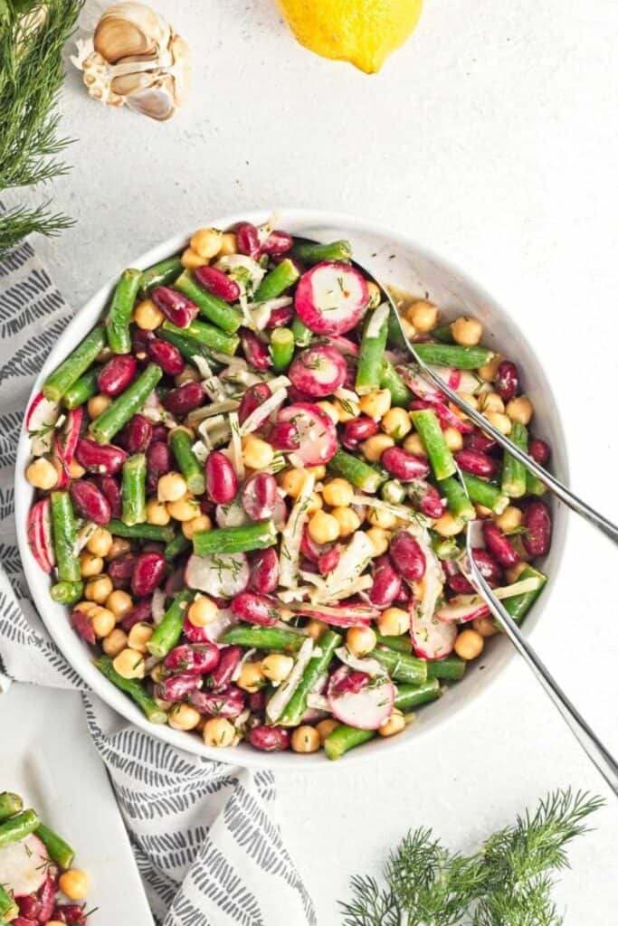Classic three bean salad in a white bowl, featuring green beans, kidney beans, and chickpeas mixed with sliced radishes, thinly sliced onions, and fresh dill. The salad is lightly dressed and served with metal utensils on a light background with a patterned tea towel.