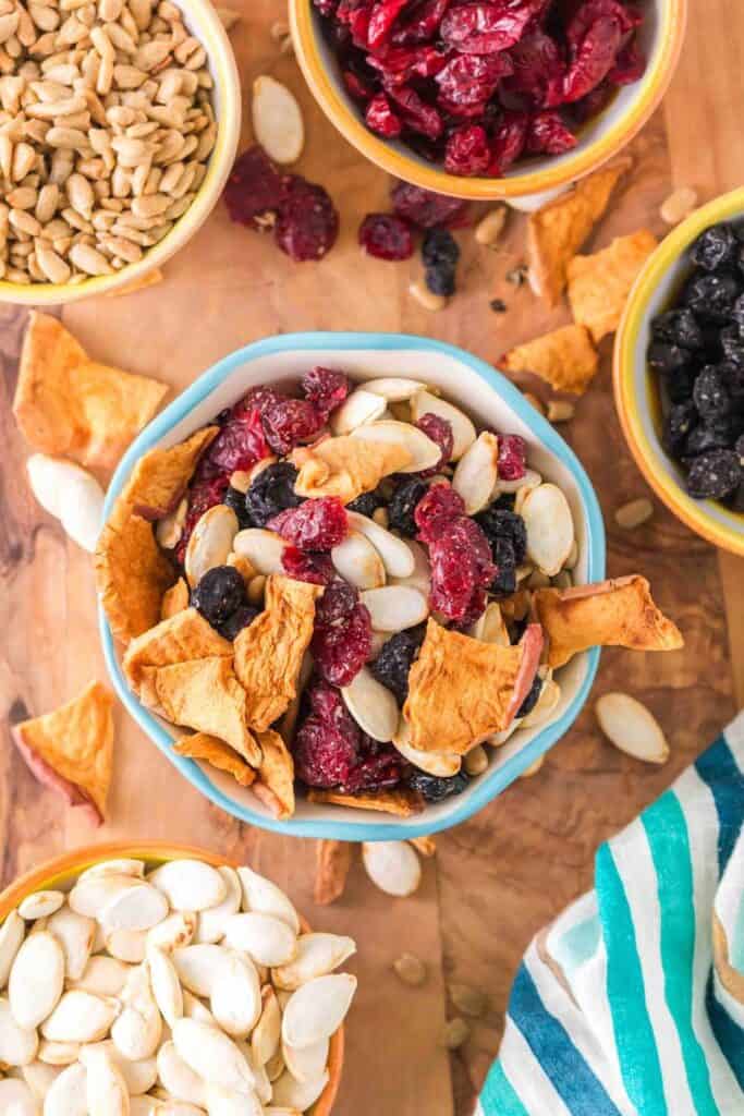 Ceramic Bowl of Coastal Medley Trail Mix, surrounded by smaller bowls of roasted sunflower seeds, and dried raisins, cranberries and pumpkin seeds. 