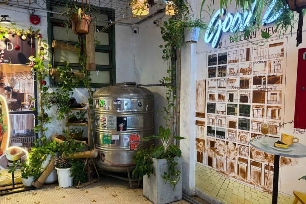 A cozy corner of a Vietnamese café is decorated with potted plants, bamboo accents, and a large metal water tank covered in colorful stickers. A hand-painted mural on the wall depicts a stylized building directory of the Coffee partments with labeled storefronts, next to a small round table holding two drinks and a dessert.