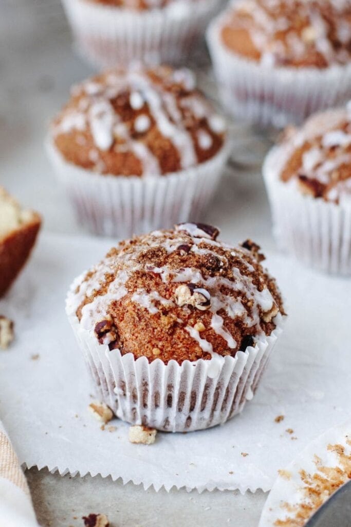Coffee cake muffins topped with crumb streusel and drizzled icing sitting in white liners on a light surface.