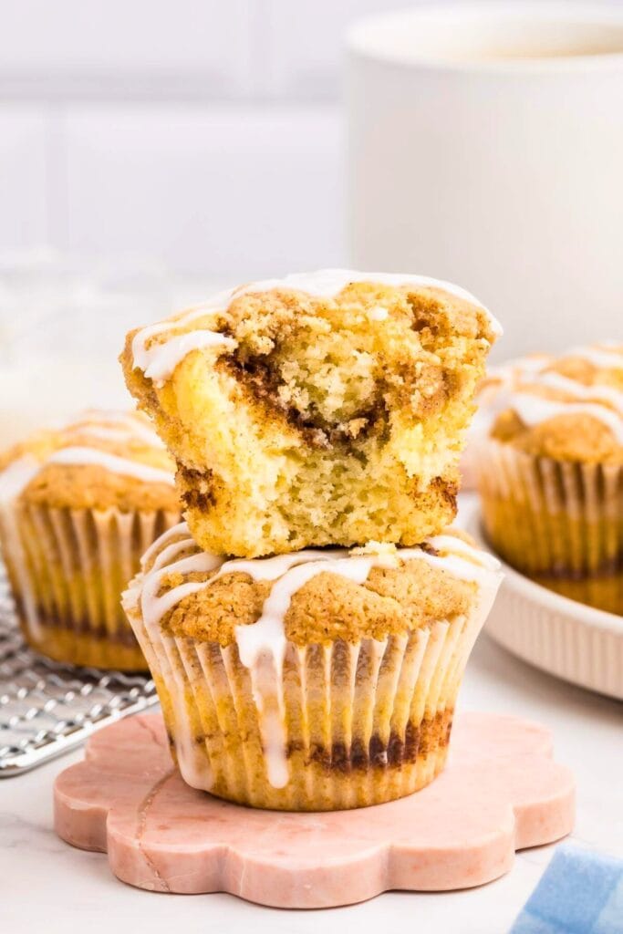 Close-up of two coffee cake muffins stacked, with the top muffin missing a bite to reveal its cinnamon-swirled interior. Both muffins are drizzled with white icing and topped with a crumbly streusel, sitting on a pink flower-shaped coaster for a cozy breakfast or snack.