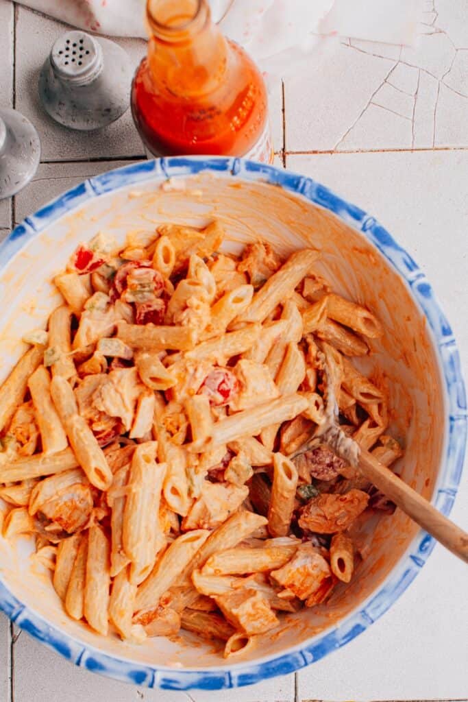 Bowl of cold Buffalo chicken pasta salad with penne pasta chunks of chicken celery and tomatoes tossed in a creamy Buffalo sauce. A bottle of hot sauce and salt and pepper shakers sit beside the bowl on a tiled table.