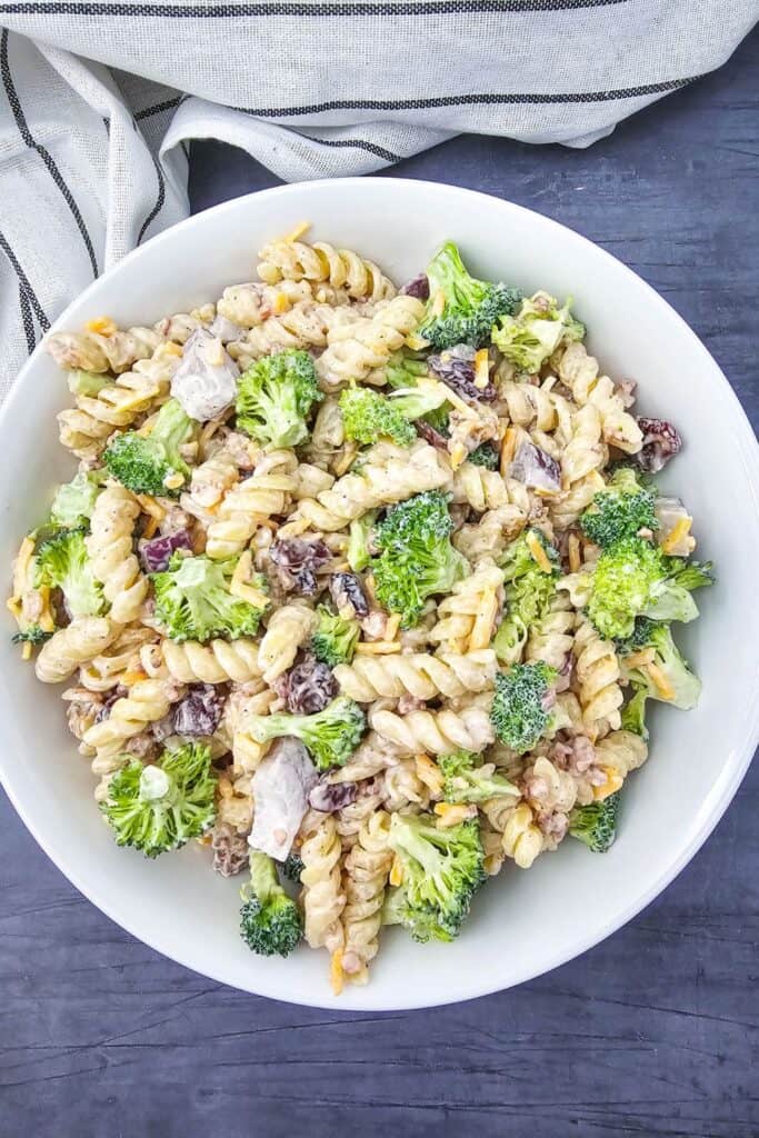 Large white bowl filled with cold creamy broccoli pasta salad, made with rotini pasta, broccoli florets, shredded cheddar, bacon bits, dried cranberries, and chunks of chicken, all coated in a thick, peppery dressing.