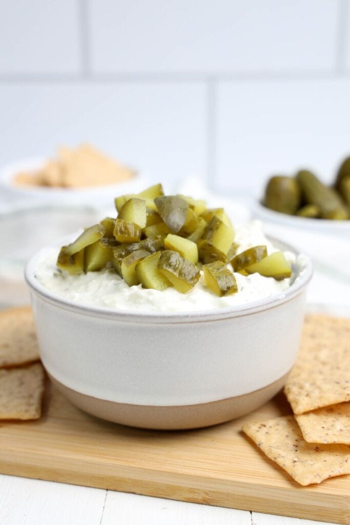 A creamy dill pickle dip topped with chopped pickles, served in a white and beige ceramic bowl. Crackers are arranged on a wooden board around the bowl, with more pickles and ingredients blurred in the background.