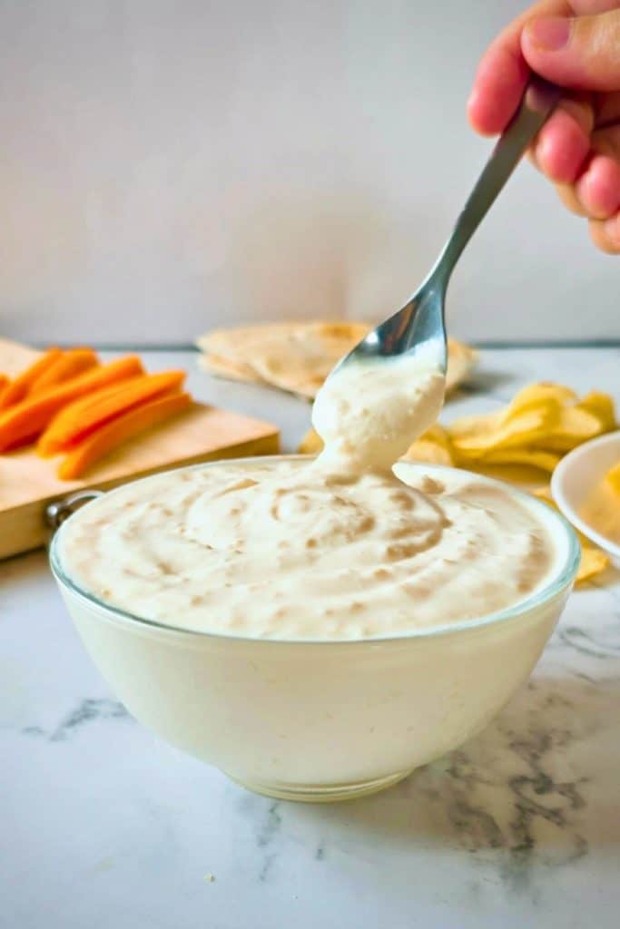 Creamy garlic dip in a glass bowl, with a spoon lifting a smooth scoop from the top. This cold tailgate dip is surrounded by chips, carrot sticks, and flatbread.