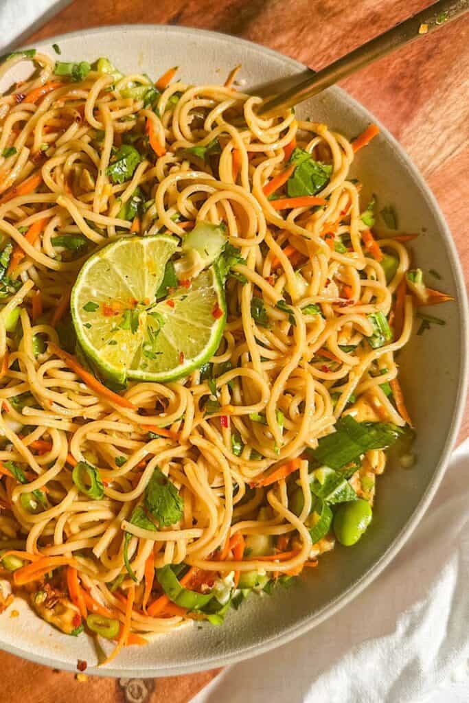 Cold ramen salad served in a shallow bowl, featuring noodles tossed with shredded carrots, chopped greens, edamame, scallions, and a soy-ginger dressing. A fresh lime slice rests on top, adding a bright citrus garnish to the vibrant, refreshing dish.