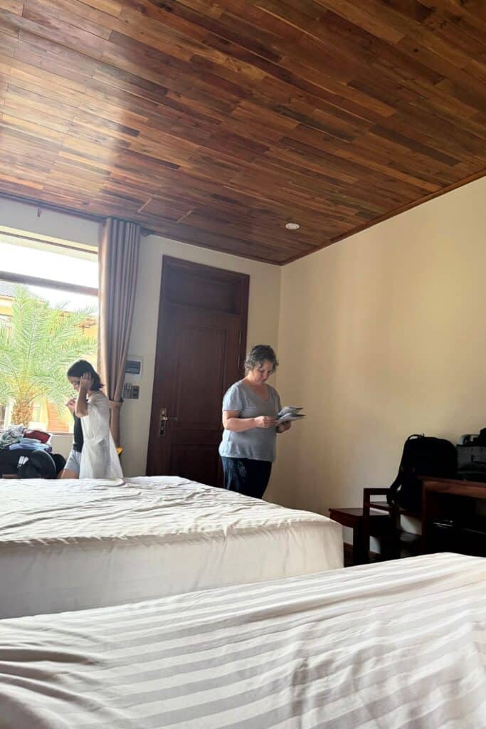 Family friendly resort room at Palm Bay Resort on Phu Quoc Island, featuring two comfortable white-striped beds, wooden ceiling, and simple furnishings. A woman reviews resort information while a child stands near the window overlooking palm trees.