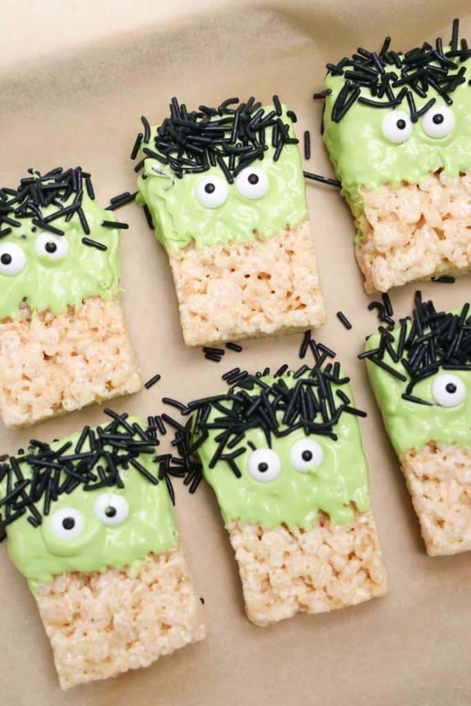 Frankenstein Rice Krispie treats in mid-decoration, with green candy-coated tops, black jimmies for hair, and candy eyes placed on each treat. The spooky snacks are arranged on parchment paper.